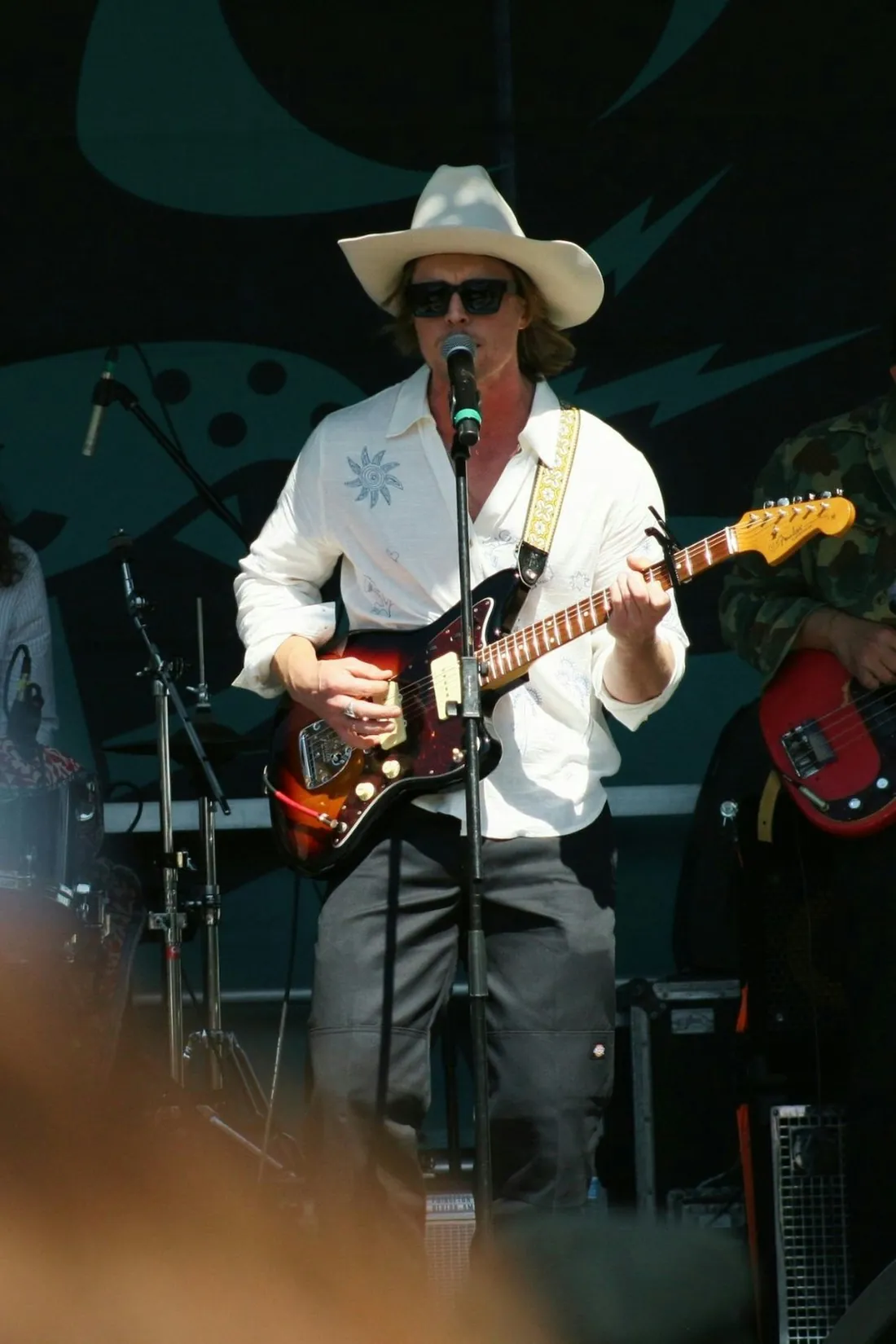 A musician in a cowboy hat singing into a mic while playing electric guitar at an outdoor festival.